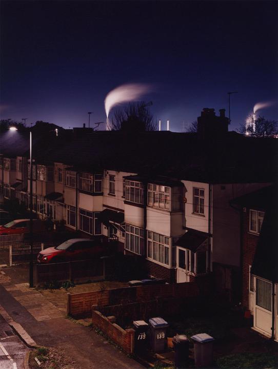 A row of houses with bay windows and parked cars is visible at night, with industrial smoke plumes rising in the distance.