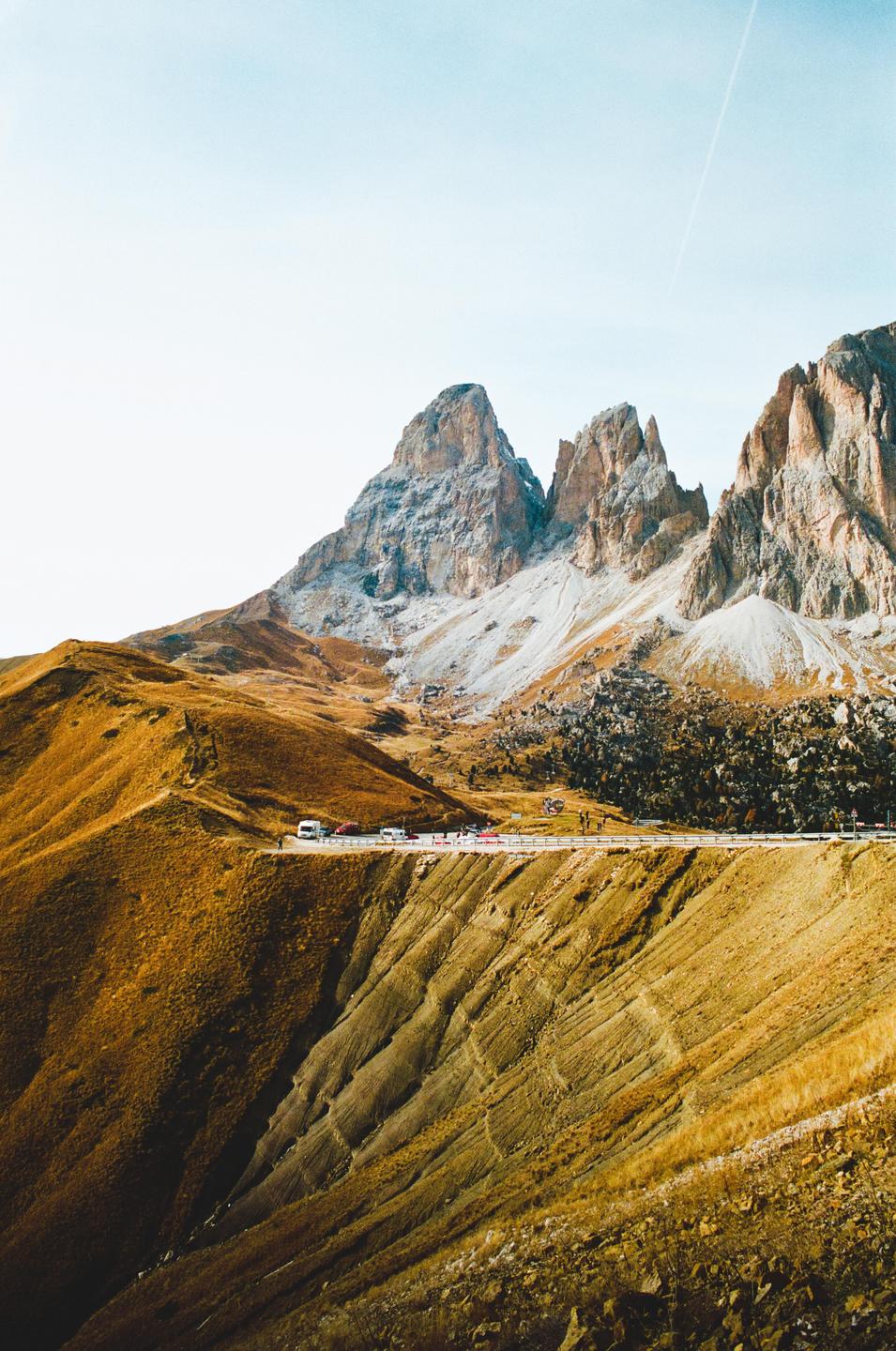 A road with cars and people winds through a mountainous landscape with rocky peaks and golden hillsides.