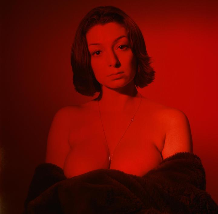 A woman with short hair and a necklace is illuminated by red light, wearing a dark, furry garment.