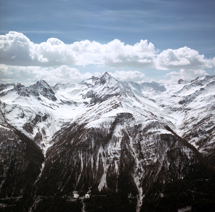 A range of snow-covered mountains with trees on their lower slopes stretches under a cloudy sky.