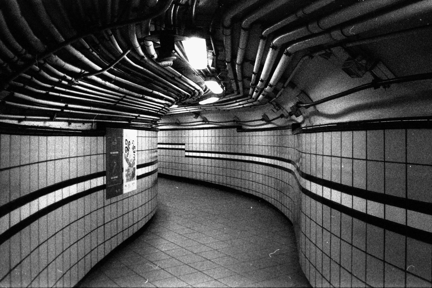 A curved, tiled subway tunnel with exposed pipes on the ceiling is illuminated by overhead lights.