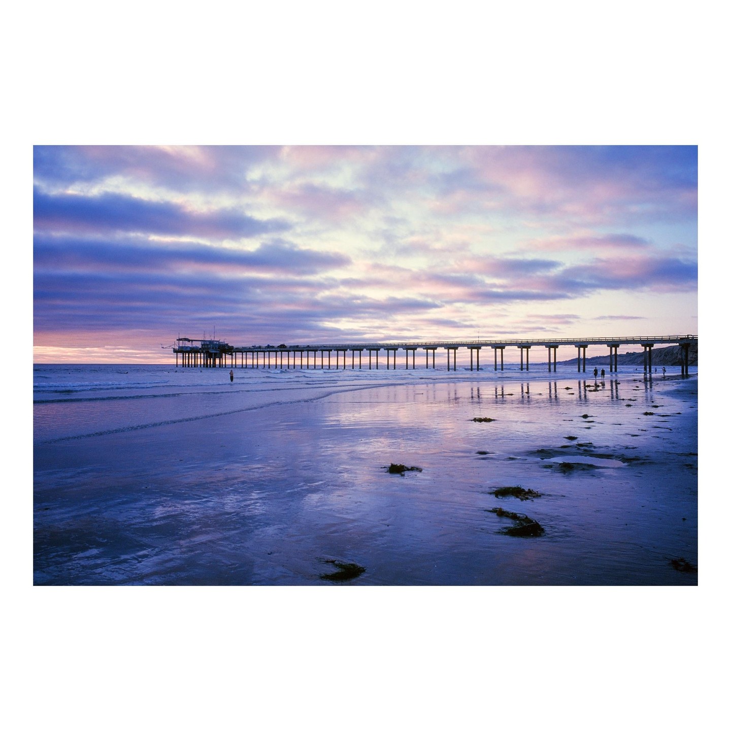 A long pier stretches into the ocean under a colorful sunset sky with reflections on the wet sand.