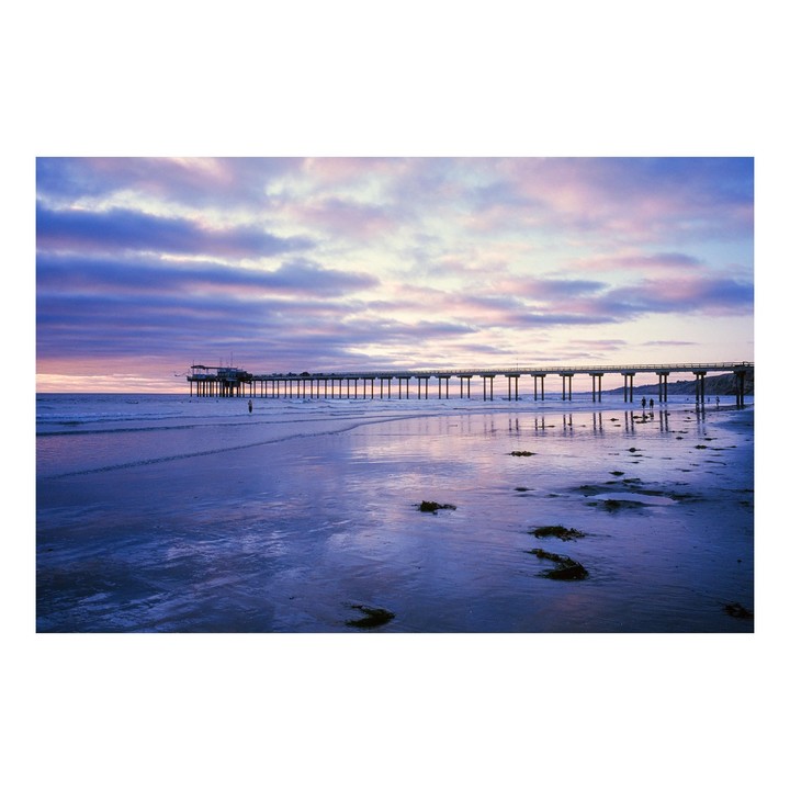 A long pier stretches into the ocean under a colorful sunset sky with reflections on the wet sand.
