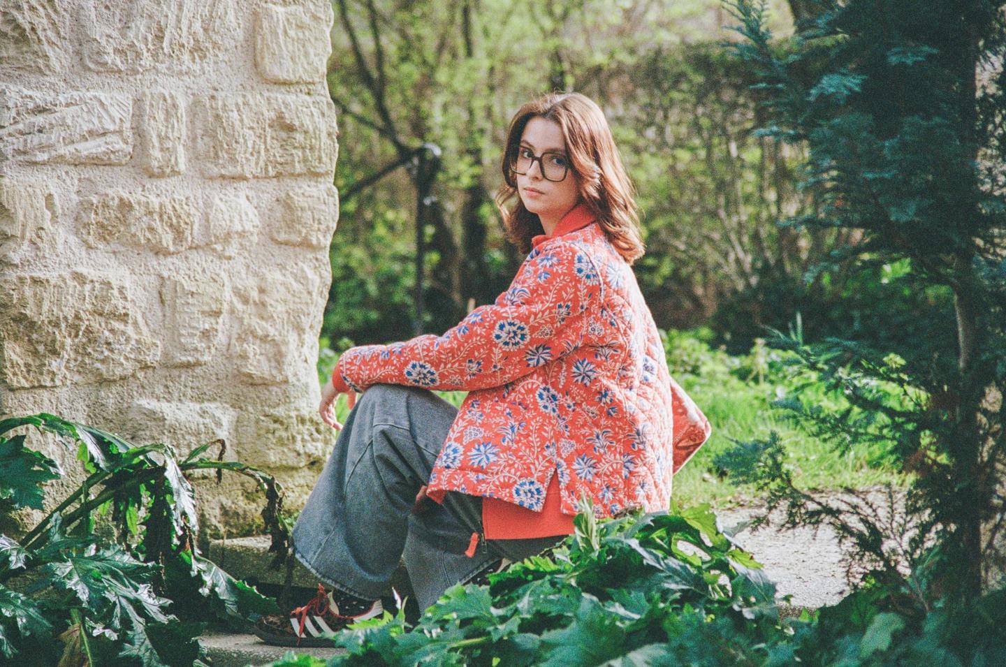 A woman wearing a patterned jacket and glasses sits on a stone step surrounded by lush green foliage.
