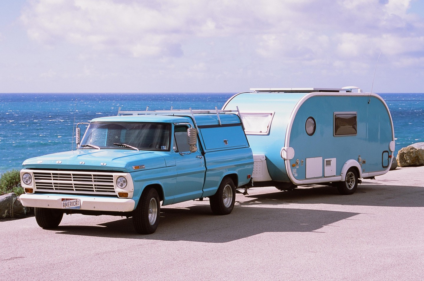 A light blue pickup truck with a matching camper shell is parked next to a light blue teardrop trailer, with the ocean visible in the background.