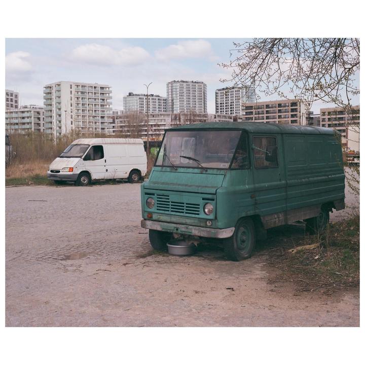 Two vans are parked on a paved lot with tall residential buildings in the background.