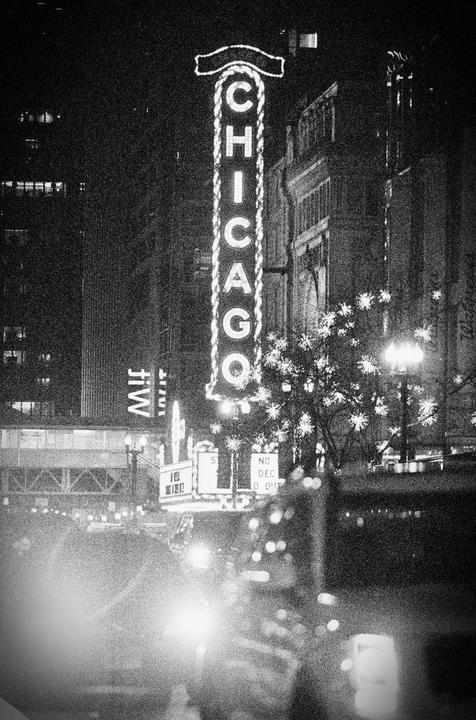 A tall neon sign spelling "CHICAGO" stands prominently at night, with blurred lights and buildings in the background.