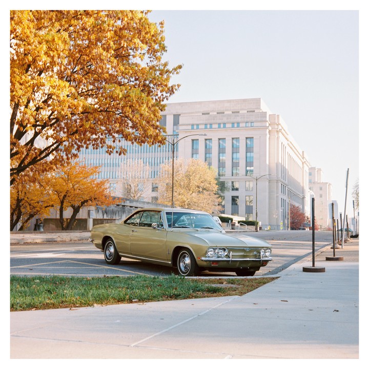 A gold vintage car is parked on a street next to a sidewalk with autumn trees and buildings in the background.