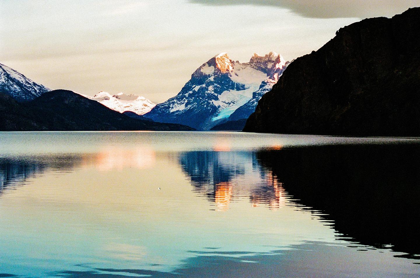 Snow-capped mountains are reflected in a calm lake under a cloudy sky.