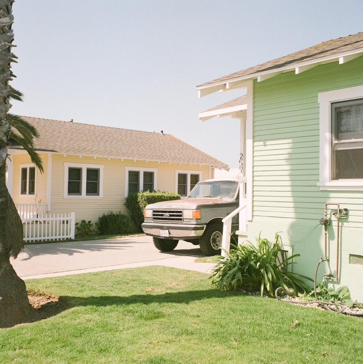 A pickup truck is parked in front of a light green house with a yellow house in the background.
