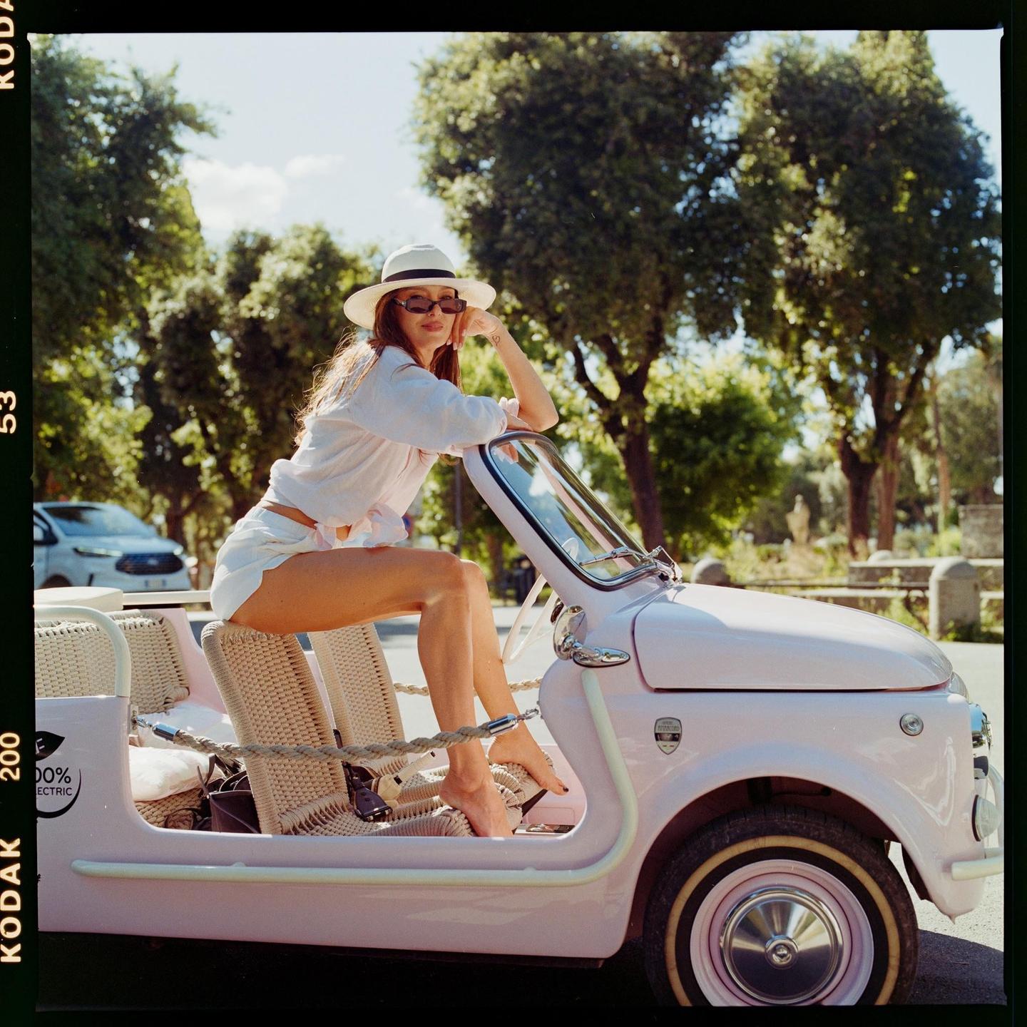 A woman wearing a hat and sunglasses is sitting in a pink car.