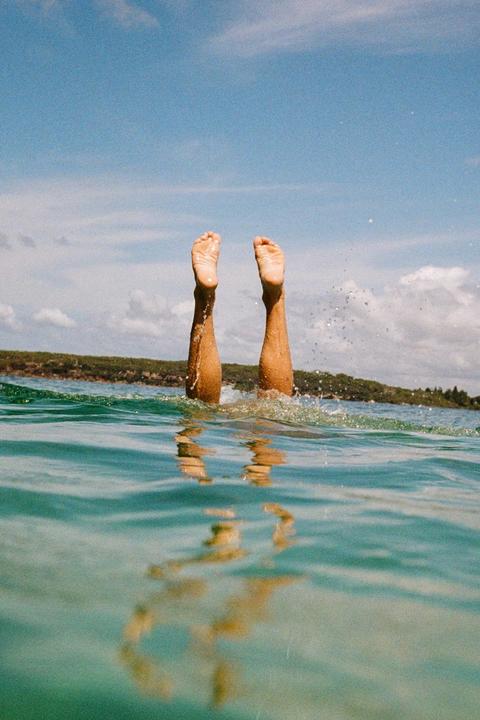 A person's legs and feet are visible above the water as they dive into a body of water under a blue sky.