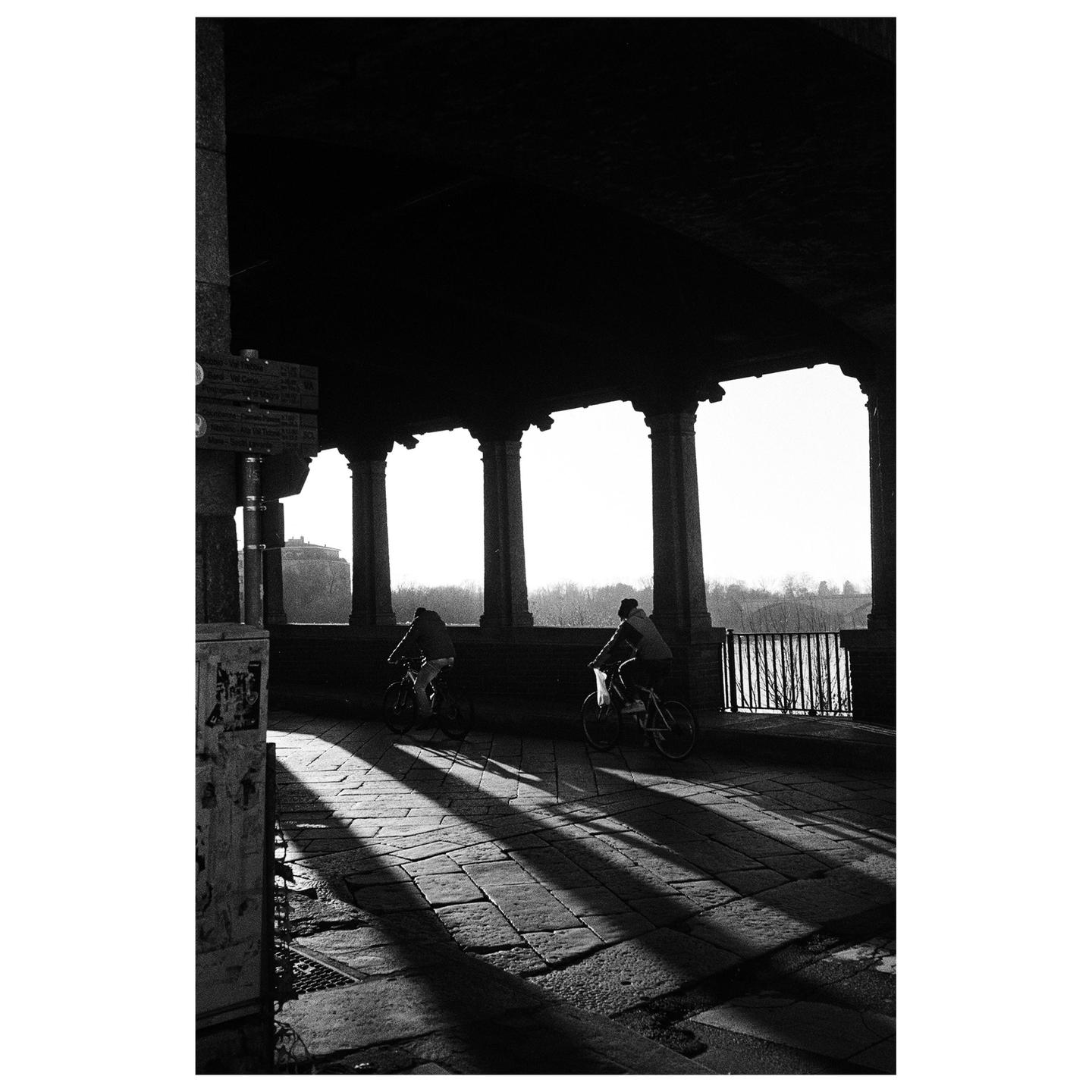 Two people ride bicycles under a stone archway with strong shadows cast on the ground.