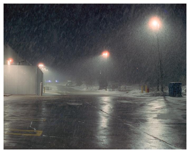 A snowy parking lot at night is illuminated by orange streetlights.