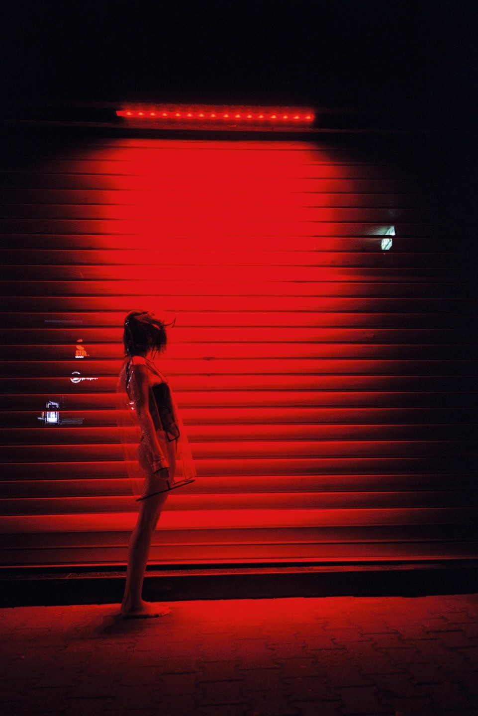 A woman stands barefoot in front of a red-lit corrugated metal wall at night.