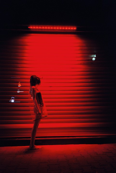 A woman stands barefoot in front of a red-lit corrugated metal wall at night.