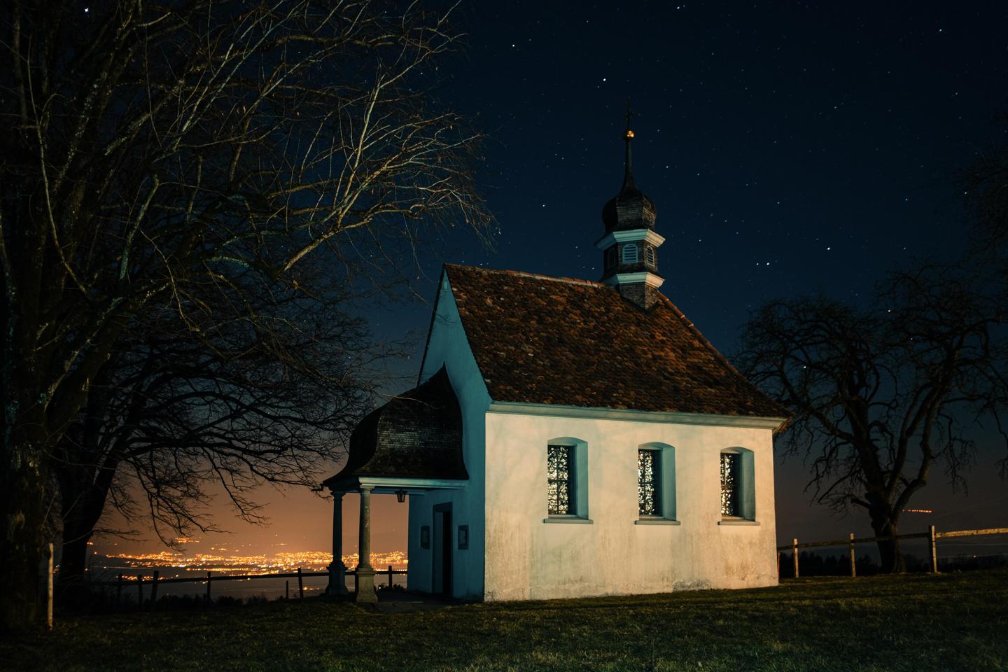 A small white chapel with a brown roof stands on a grassy hill at night, with a city glowing in the distance and stars visible in the dark sky.