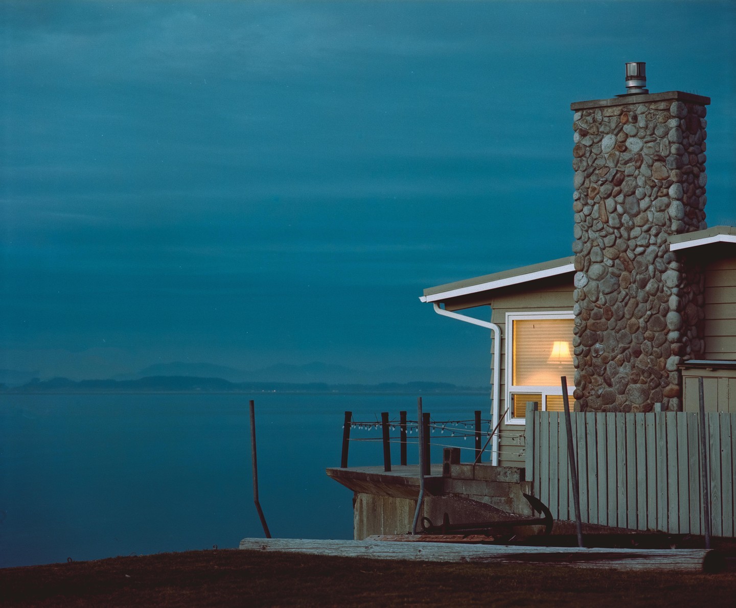 A house with a stone chimney overlooks a body of water with mountains in the distance at dusk.