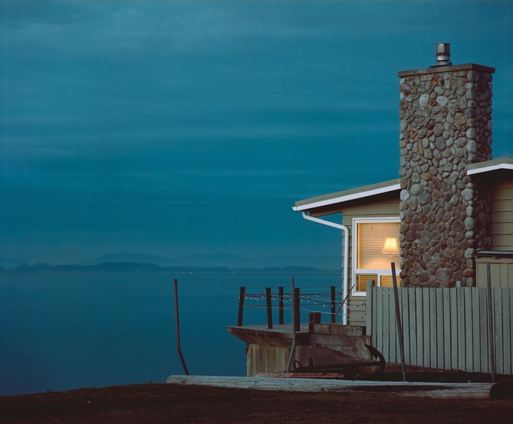 A house with a stone chimney overlooks a body of water with mountains in the distance at dusk.