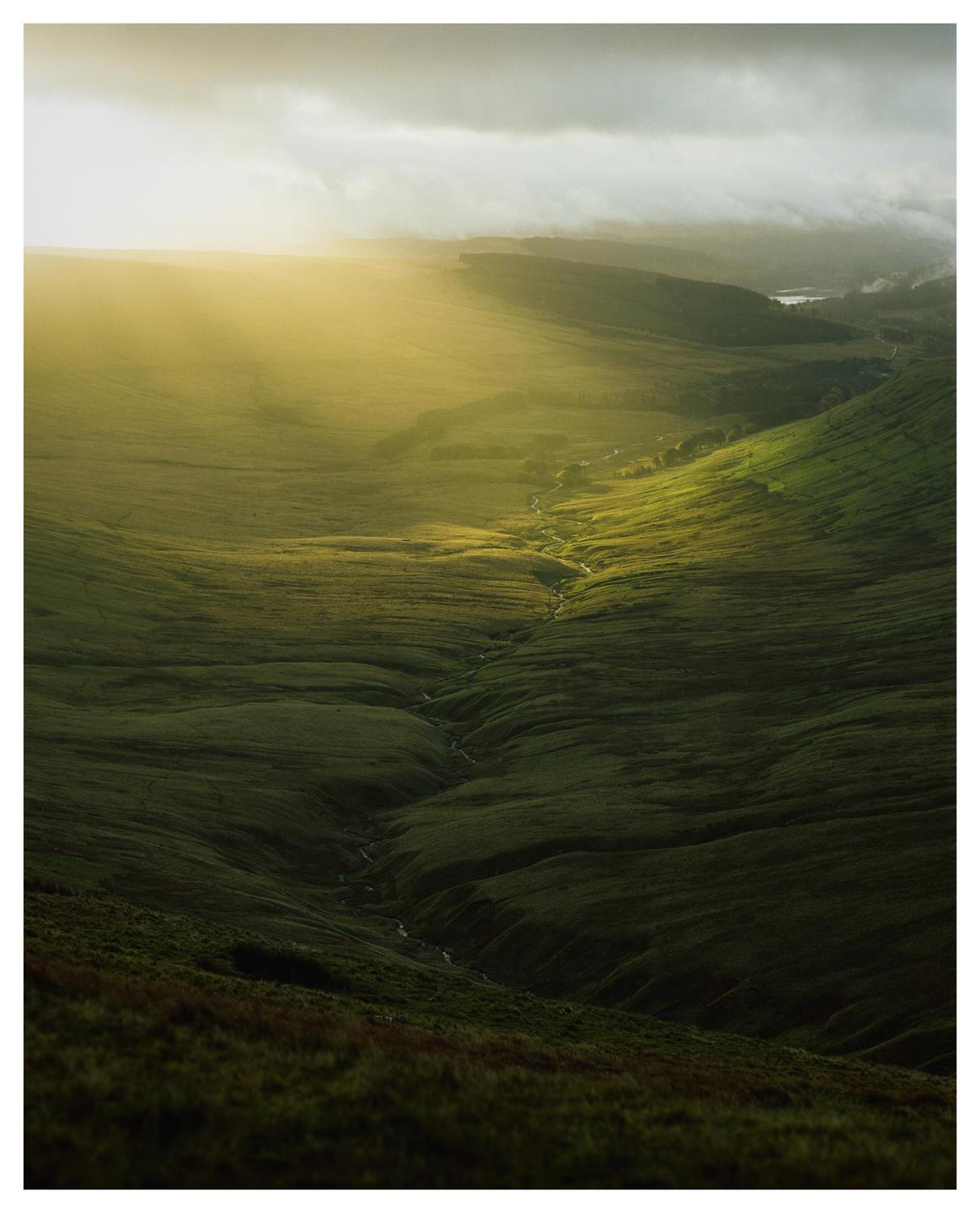 Sunlight breaks through clouds over a valley with a winding stream.