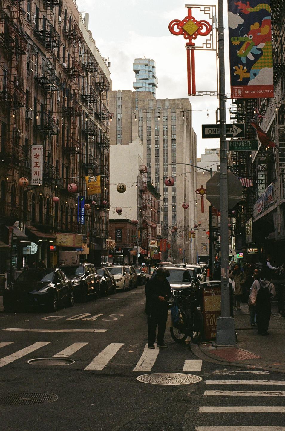 A street in Chinatown, New York, is filled with cars, pedestrians, and buildings adorned with fire escapes and lanterns.