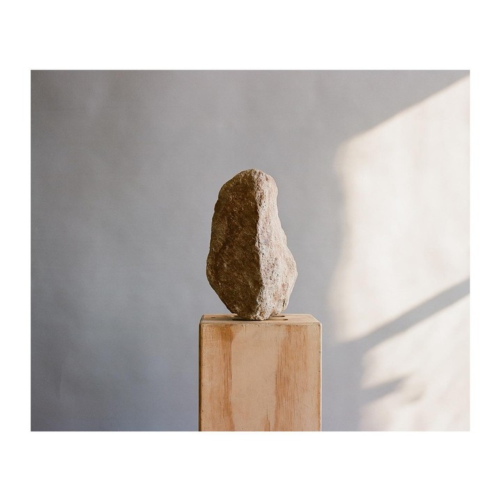 A tall, rough-hewn stone stands on a wooden pedestal against a plain wall with a shadow cast on it.