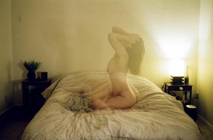 A translucent woman is captured in a self-portrait, kneeling on a bed in a dimly lit room.