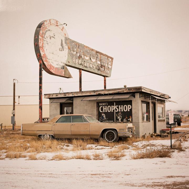 A vintage car is parked in front of a small building with a large, weathered sign that reads "Gasoline Refinery" and "Rachet's Chop Shop."