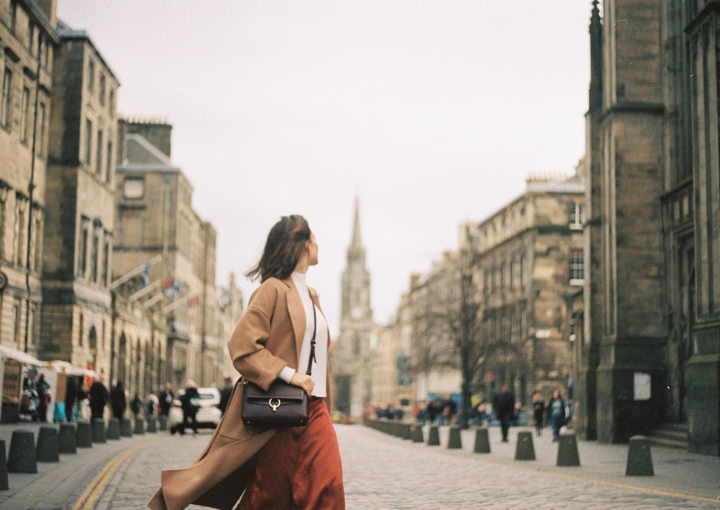 A woman in a long coat and skirt walks down a city street with historic buildings and a church steeple in the background.