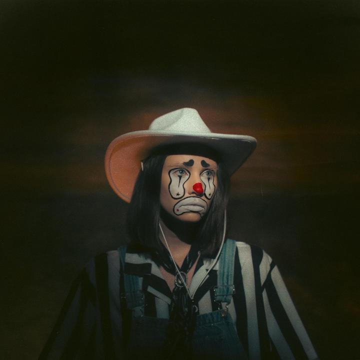 A woman wearing a cowboy hat and clown makeup looks up and to the right.