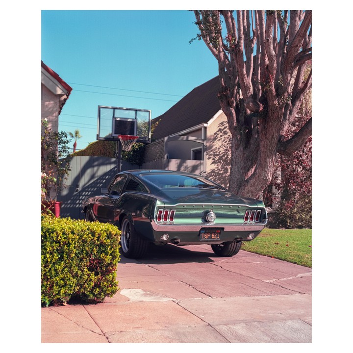 A green vintage car is parked in a driveway next to a basketball hoop and a large tree.