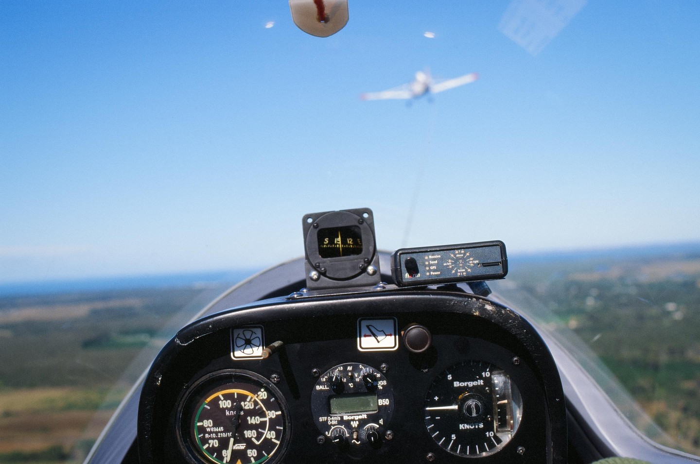 The cockpit of a glider is visible with a small plane in the distance against a clear blue sky.