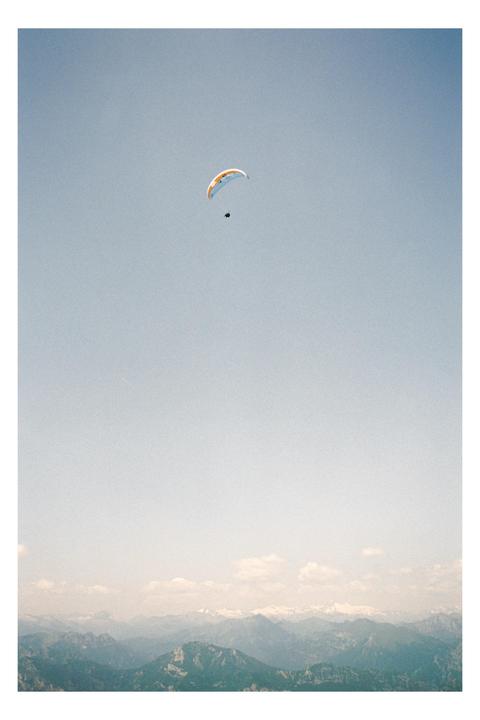 A paraglider is visible in the sky above a mountain range with some snow-capped peaks.