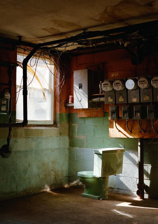 Sunlight streams through a window into a basement room with a green toilet, exposed wiring, and utility meters.