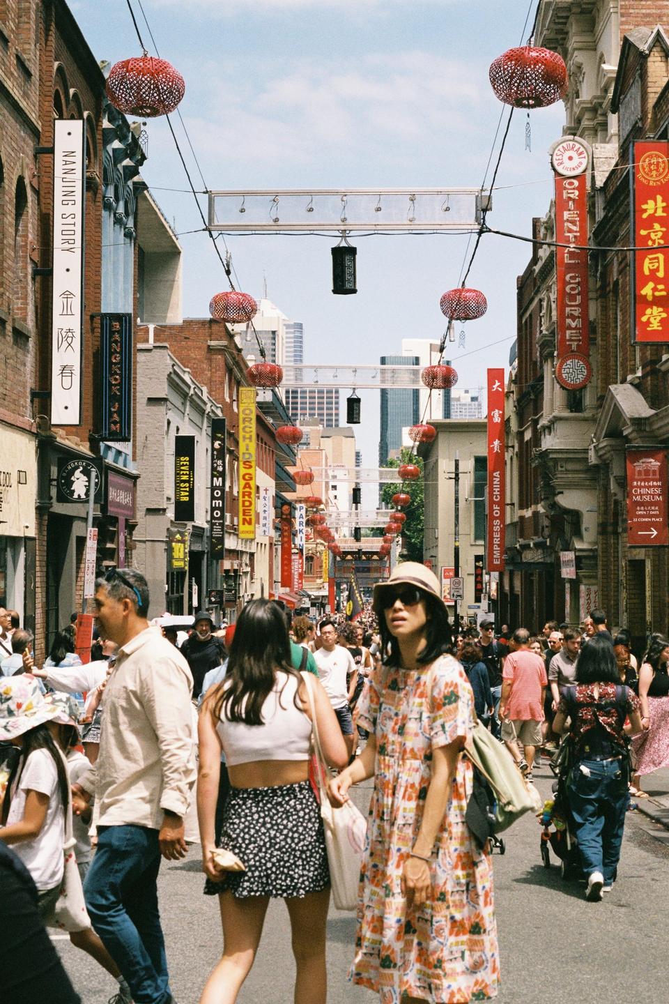 A street is filled with people walking past shops and red lanterns hanging overhead.