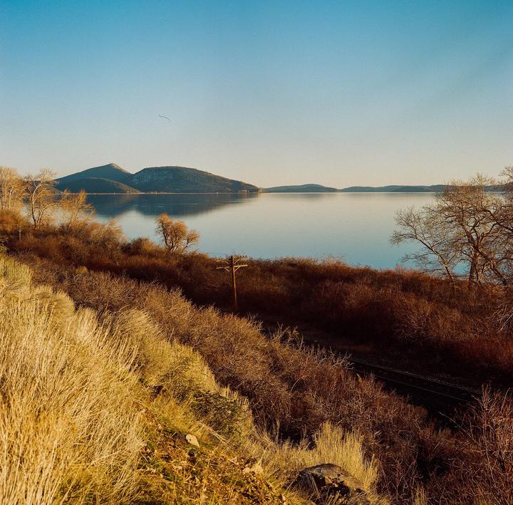 A body of water reflects mountains under a clear sky, with dry brush and a telephone pole in the foreground.