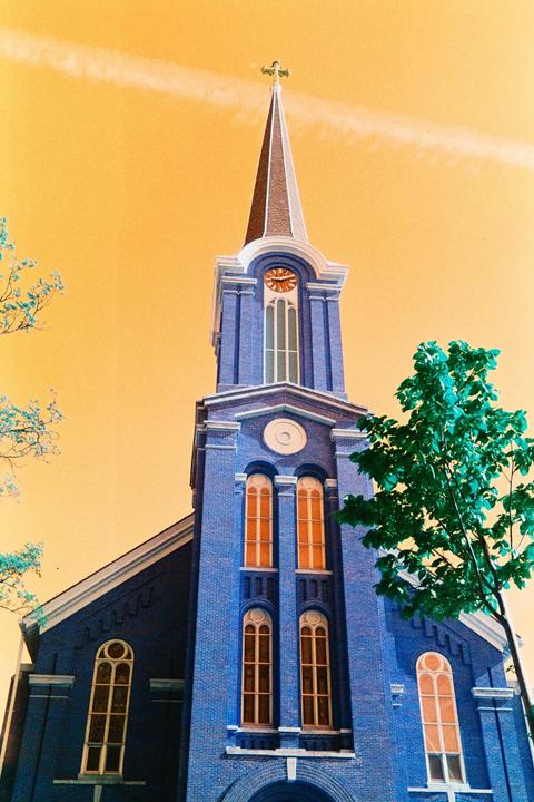 A blue brick church with a tall steeple and clock tower stands against an orange sky with a tree in the foreground.