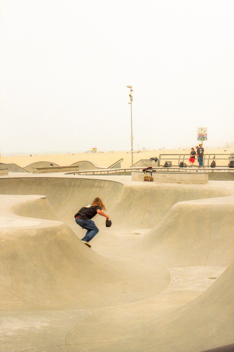 A person with long hair skates in a concrete skatepark with a beach and ocean in the background.