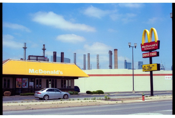 A McDonald's restaurant with a silver car parked in front of it, and industrial smokestacks visible in the background under a blue sky.