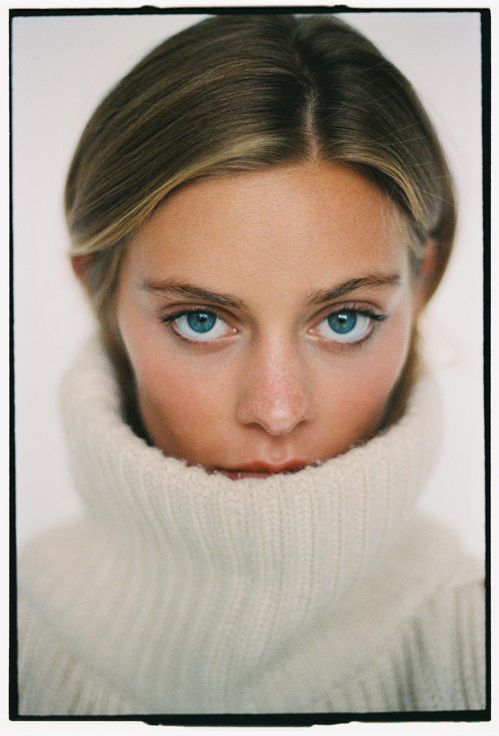 A woman with blue eyes looks directly at the viewer, her face partially obscured by a cream turtleneck sweater.