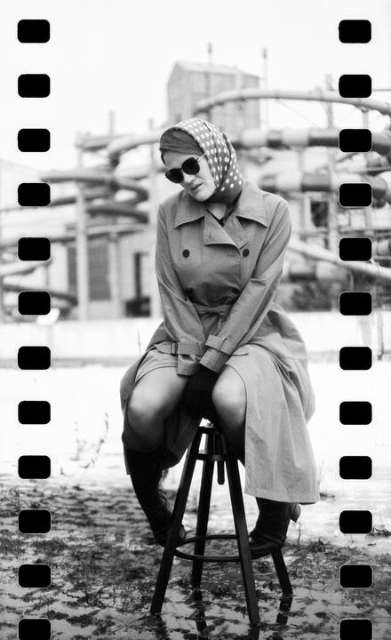 A woman wearing a trench coat, headscarf, and sunglasses sits on a stool in a flooded area with industrial structures in the background.