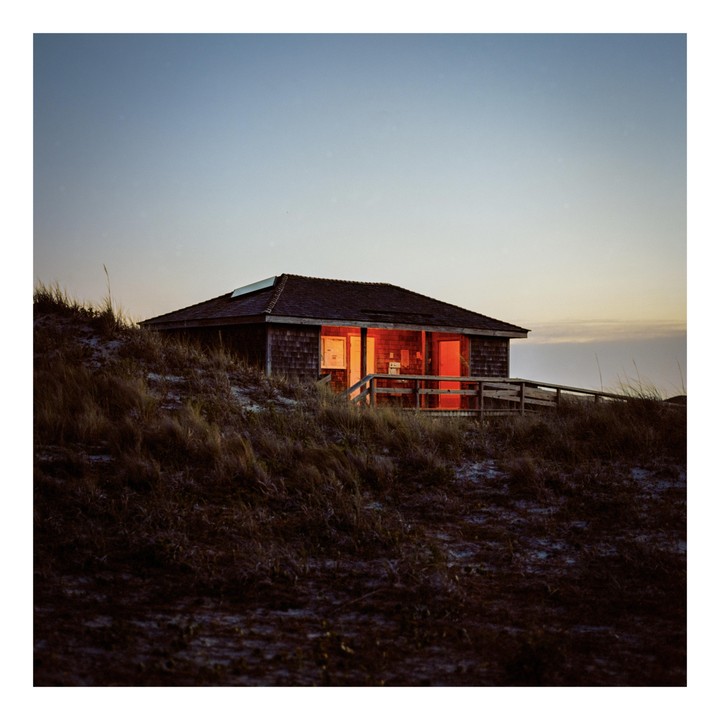 A small house with glowing orange windows sits on a grassy hill under a pale sky.