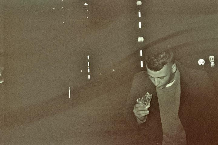 A man in a coat and sweater holds a piece of pizza at night.