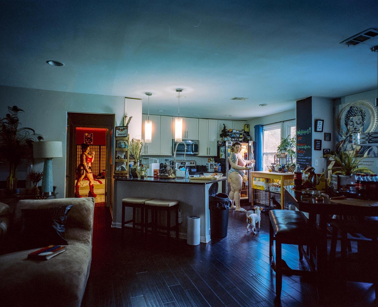 A woman in a red outfit stands in a doorway while another woman stands by an open refrigerator in a kitchen with a small dog.
