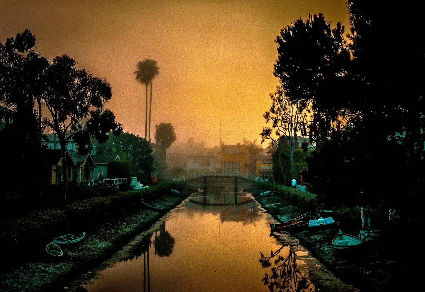 A canal with a bridge and houses on its banks is shrouded in fog at sunset.