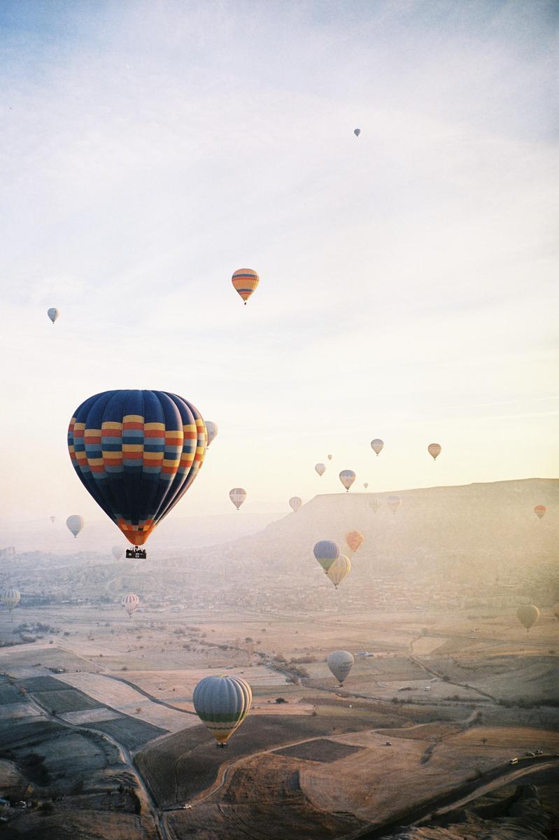 Many hot air balloons float above a hazy, mountainous landscape at dawn.
