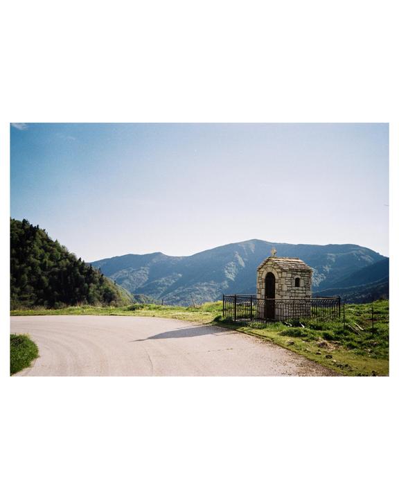 A small stone chapel with a cross on its roof stands beside a winding road, with mountains and trees in the background.