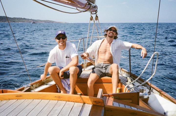 Two men are sitting on a sailboat on the ocean with an island in the distance.