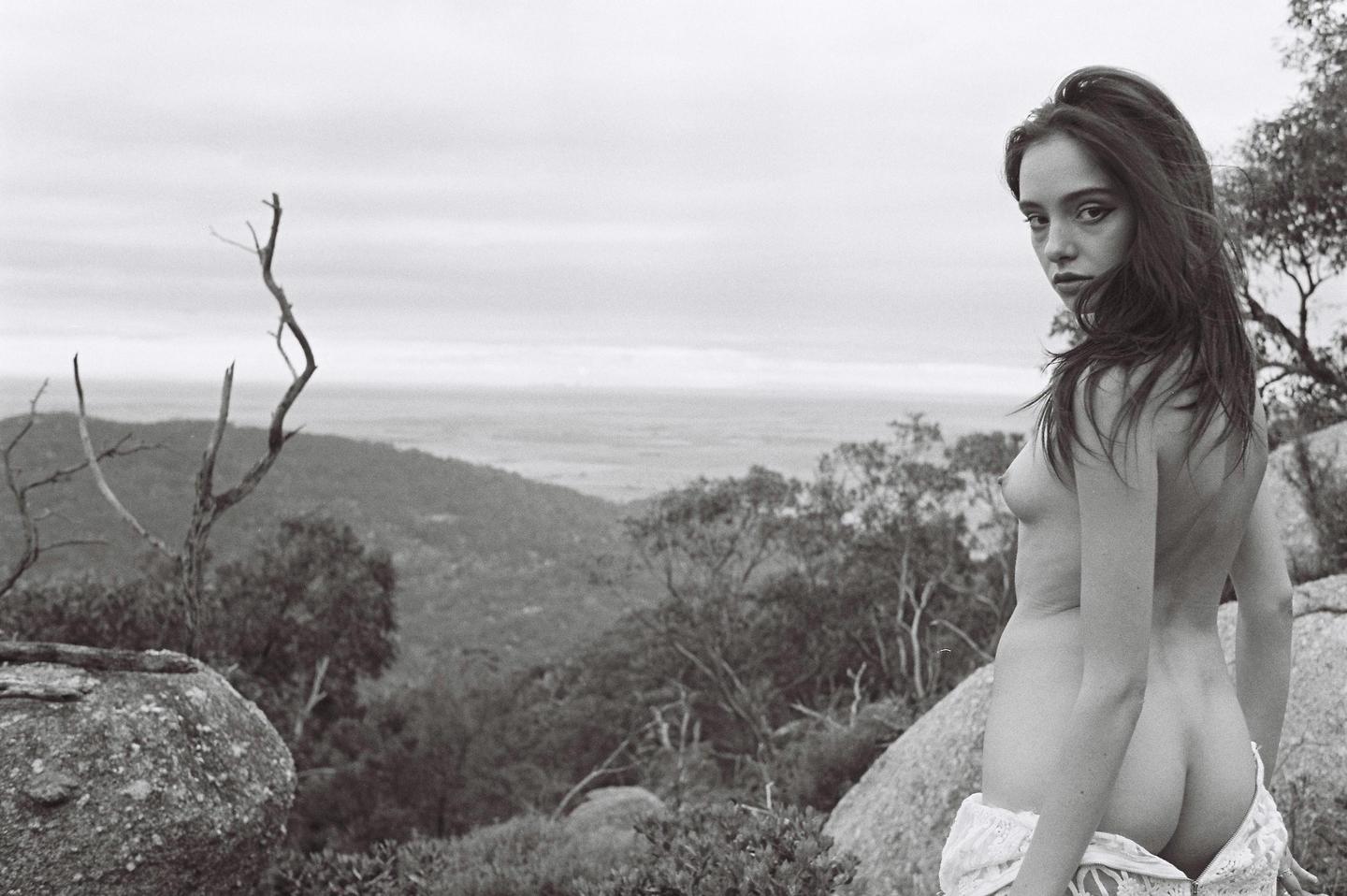 A woman stands topless on a rocky hillside overlooking a valley.