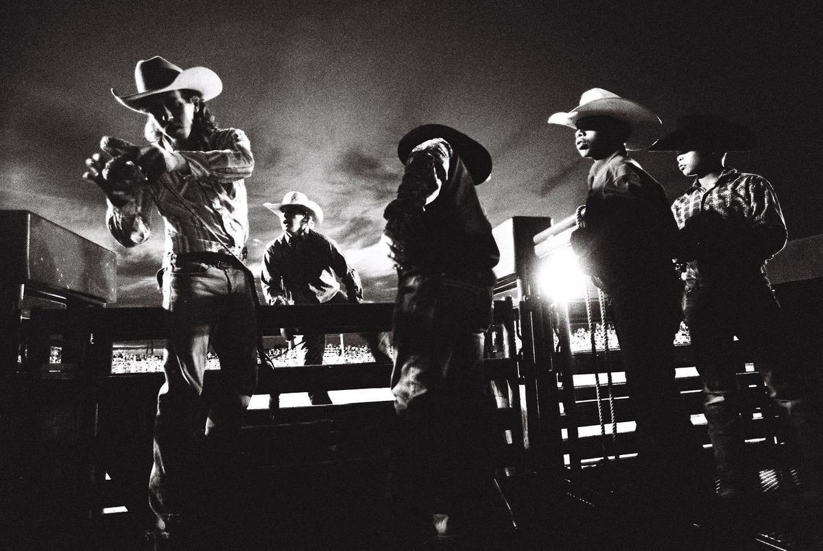 Several men in cowboy hats are standing on a wooden structure at night, with one man in the foreground adjusting something in his hands.